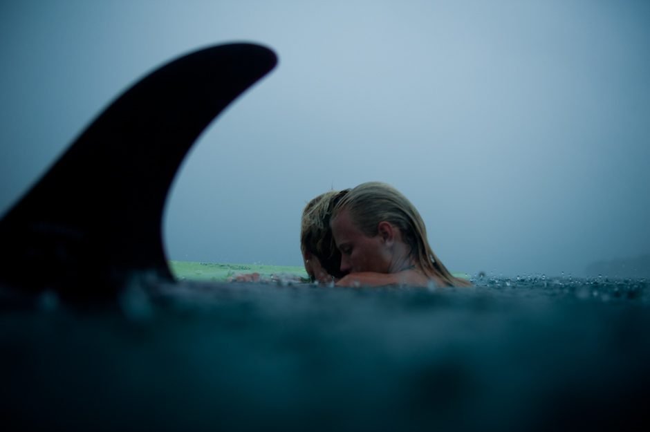 An image of a male and female surfer rest in the water beside a surfboard