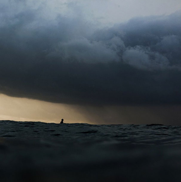 An image of a surfer sits on his board under a dark storm cloud