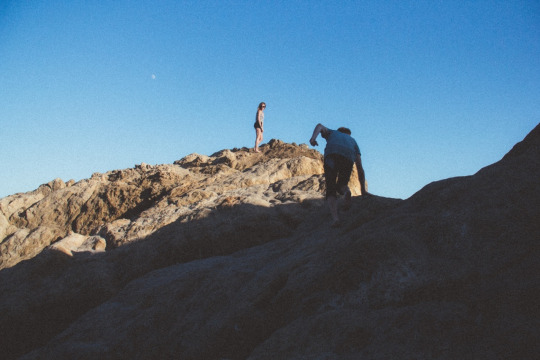 An image of a man scrambling up a ridgeline to a woman waiting above
