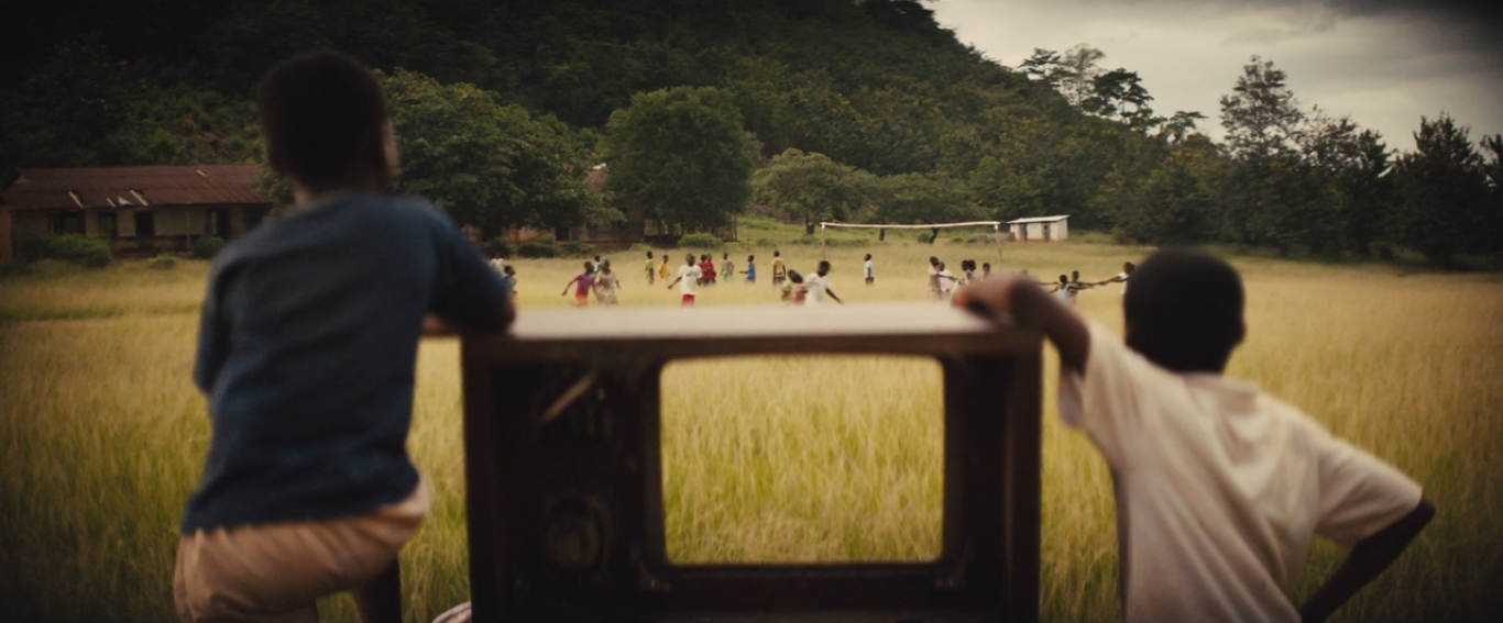 Two boys in the foreground lean against an old TV while watching others play in a still from the film Beasts of No Nation