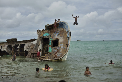 A boy leaps off the bow of a wrecked freighter while other children play in the water below in this image by Kadir van Lohuizen