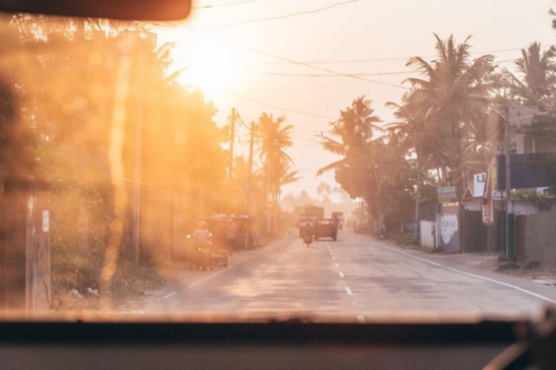Looking out the window of a vehicle towards a palm-lined road with a Tuk Tuk framed by a setting sun
