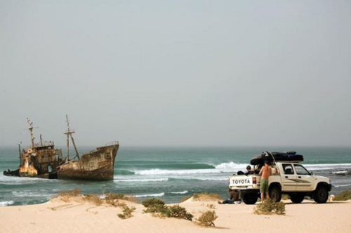A surfer stands outside of his truck, which is parked on a dune overlooking a shipwreck