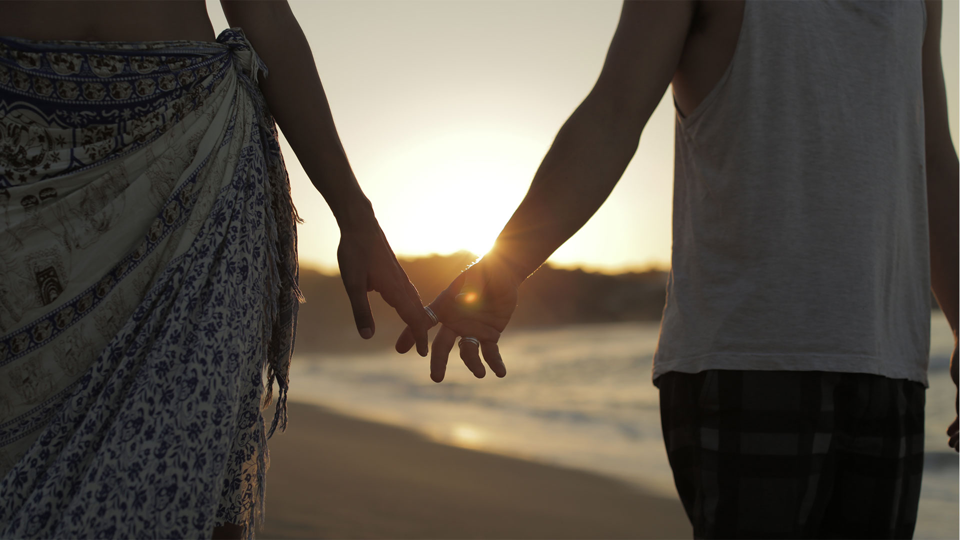 a close up of two people holding hands on a beach, backlit by a setting sun